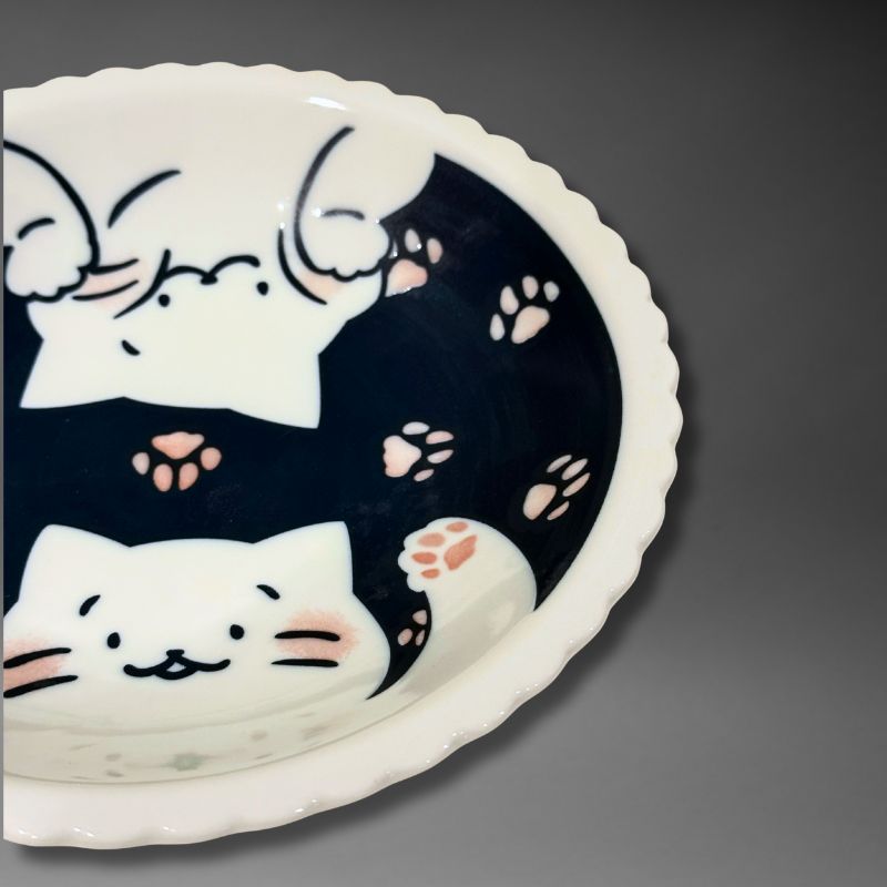 A close-up detail shot of the ceramic plate's interior, showing the face of one smiling white cat and multiple pink and brown paw prints near the scalloped rim.