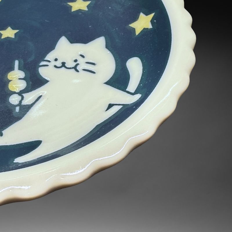 A close-up detail shot of the small ceramic plate, showing the white cat design, the yellow dango skewer, and the white, scalloped 'biscuit' rim.