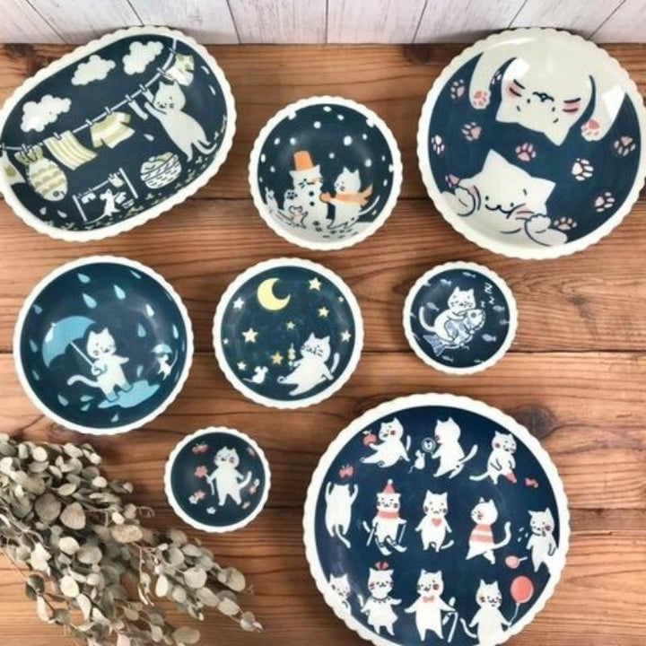 A flat lay shot showcasing the full K Factory Cat collection, featuring plates and bowls in various sizes and shapes, each with a different whimsical cat design on a dark blue background.
