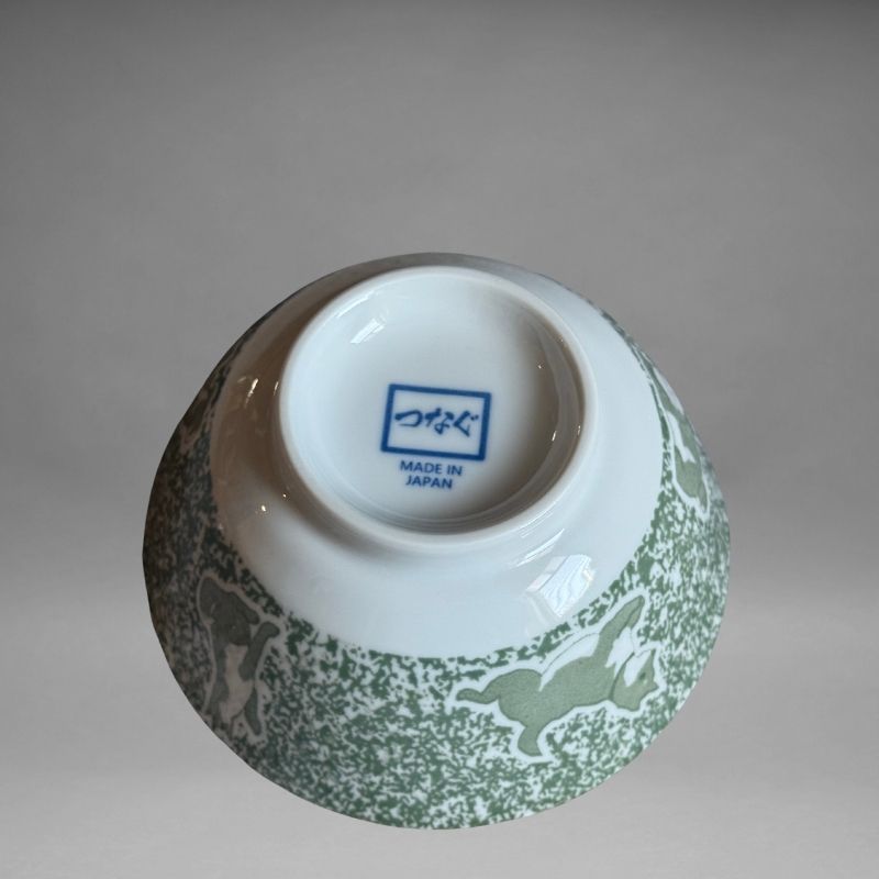 An inverted, bottom-up view of the small ceramic rice bowl, showing the white base and the blue 'つなぐ MADE IN JAPAN' mark stamped in the centre. The green/grey speckled exterior with Shiba dog designs is also visible.