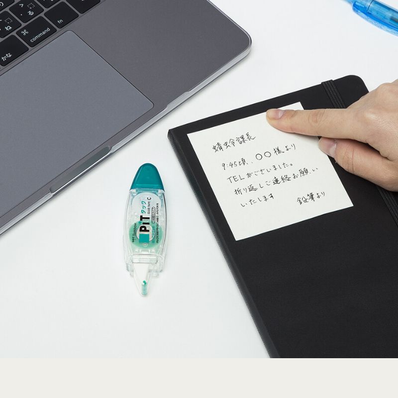 Lifestyle shot showing a handwritten note being stuck onto a black planner like a sticky note. The Tombow PiT Tack-C dispenser sits on the desk next to a laptop, demonstrating its use for creating custom removable memos.