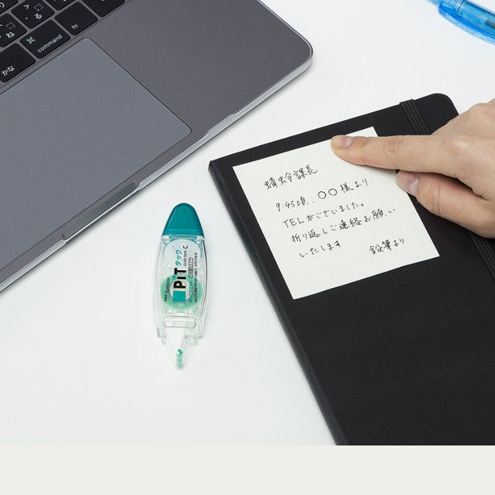 Lifestyle shot showing a handwritten note being stuck onto a black planner like a sticky note. The Tombow PiT Tack-C dispenser sits on the desk next to a laptop, demonstrating its use for creating custom removable memos.