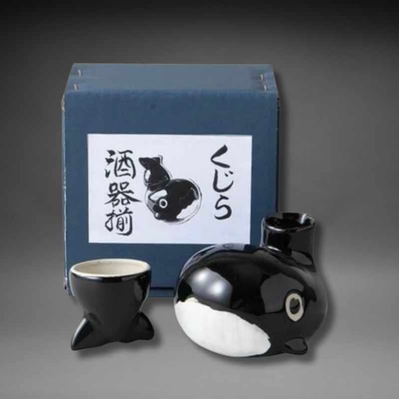 Composition photograph showing the complete dark blue Mino-yaki Whale Sake Set gift box, positioned directly behind the black and white ceramic whale-shaped tokkuri (sake decanter) and the separate black ceramic tail cup, on a simple light grey studio background. This image highlights the set as a premium gift product.