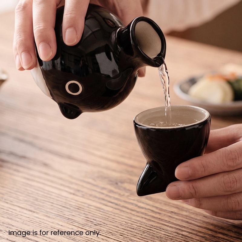 Close-up action photograph of a hand with a simple ring pouring clear Japanese sake from the black ceramic whale-shaped tokkuri (sake decanter) into the matching black ceramic tail-shaped cup