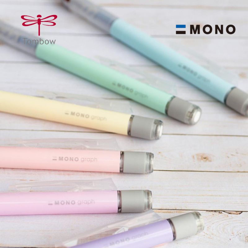 A close-up, aesthetic arrangement of Tombow Mono Graph Shaker mechanical pencils in five pastel colors—Ice Blue, Mint Green, Cream Yellow, Coral Pink, and Lavender—on a light weathered wooden surface, with "MONO graph" visible on the yellow pencil barrel.