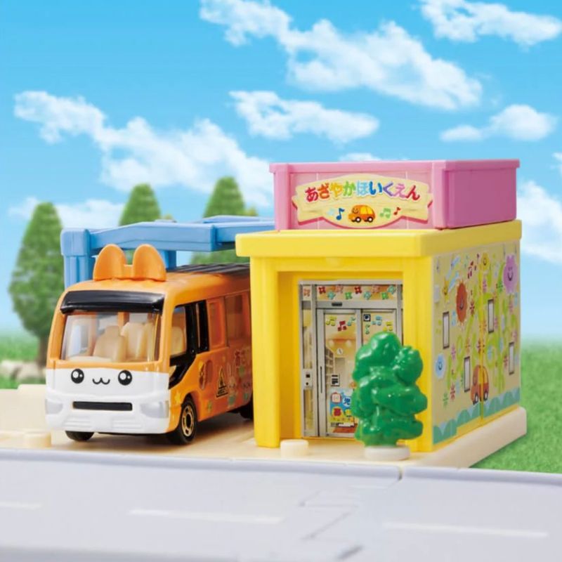 Tomica orange school bus parked at the colourful nursery building with trees and blue sky background.